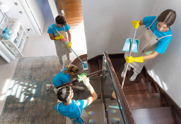 Von oben von professionellen weiblichen und männlichen Haushälterinnen in Uniformen und Gummihandschuhen, die auf einer Holztreppe stehen, während sie ein modernes geräumiges Haus reinigen - ADSF32189
