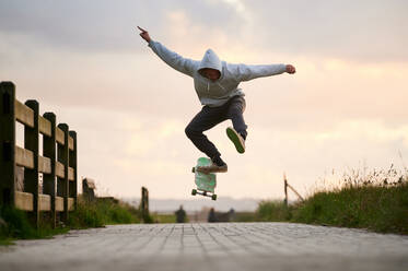 Ebenerdige Ganzkörperaufnahme eines Mannes in Freizeitkleidung, der einen Stunt auf einem Longboard auf einem gepflasterten Gehweg vollführt - ADSF32242