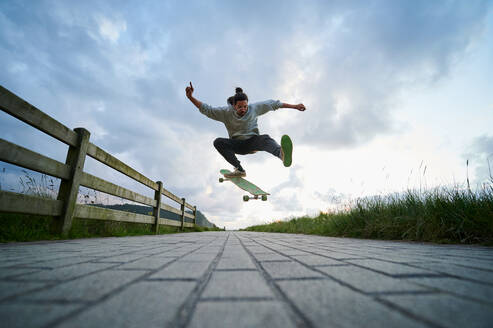 Ebenerdige Ganzkörperaufnahme eines Mannes in Freizeitkleidung, der einen Stunt auf einem Longboard auf einem gepflasterten Gehweg vollführt - ADSF32244