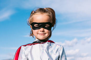 Cute kid wearing masquerade superhero costume and eye mask standing against blue sky and looking at camera - ADSF32245