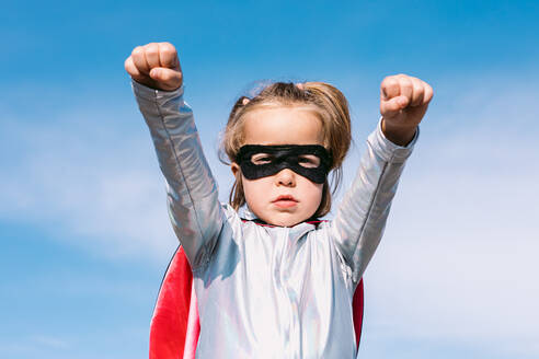 From below small girl in superhero costume raising outstretched fists for showing power while standing against blue clear sky - ADSF32247