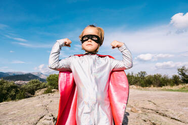 Strong girl in superhero cape raising hands for showing strength for protecting world while standing on hill and looking away with confidence - ADSF32249