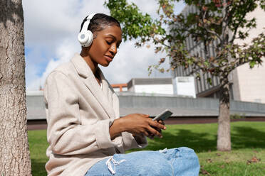 Ruhige afroamerikanische Frau mit geschlossenen Augen, die mit kabellosen Kopfhörern Musik hört, während sie auf dem Rasen in der Nähe eines Baumstamms in einem sonnigen Park sitzt und ihr Smartphone benutzt - ADSF32257