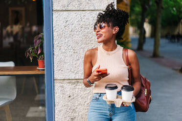 Trendy jungen ethnischen weiblichen Millennials mit dunklen Afro Haare in stilvollem Outfit und Rucksack mit Handy, während auf der Straße der Stadt mit Tassen Kaffee zum Mitnehmen stehen - ADSF32265