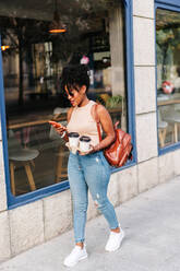 Ganzkörperaufnahme einer trendigen jungen ethnischen Frau mit dunklem Afro-Haar in stilvollem Outfit und Rucksack, die ein Mobiltelefon benutzt, während sie mit einer Tasse Kaffee zum Mitnehmen durch die Stadt läuft - ADSF32266