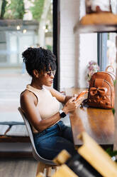 Seitenansicht einer selbstbewussten jungen afroamerikanischen Millennials mit dunklen lockigen Haaren in trendigem Outfit und Sonnenbrille, die an einem Tisch in einem modernen Café sitzt und auf ihrem Smartphone Nachrichten schreibt - ADSF32267