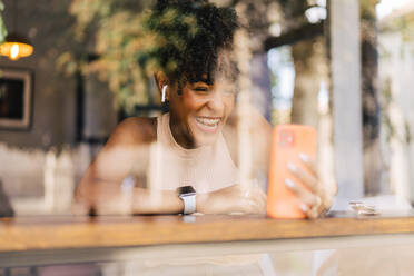 Durch das Fenster einer fröhlichen jungen ethnischen Frau mit dunklem Afro-Haar in trendigem Outfit und echten kabellosen Kopfhörern, die glücklich lächelt, während sie ein Videogespräch über ein Smartphone in einem modernen Café führt - ADSF32269