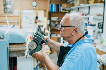 Side view of concentrated senior male carpenter with white hair in apron and eyeglasses cutting wooden boards with circular saw during work in joinery - ADSF32273