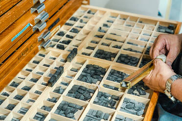 Von oben konzentrierter älterer männlicher Handwerker in Schürze und mit Brille, der während der Arbeit in einem traditionellen Atelier Druckbuchstaben aus einem Holzkasten auswählt - ADSF32276