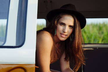 Attraktives brünettes Mädchen mit Hut in einem Oldtimer-Van, das auf dem Sitz sitzt und in die Kamera schaut, an einem sonnigen Tag - ADSF32295