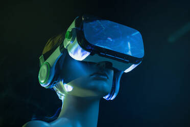 Dummy of woman in futuristic VR goggles placed under bright projection in dim room - ADSF32298