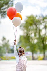 Fröhliches afroamerikanisches Mädchen mit Zöpfen in stilvoller Kleidung läuft mit bunten Luftballons in der Hand im Park am Tag - ADSF32317