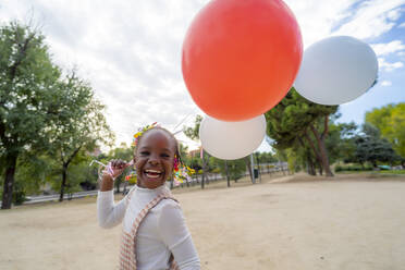 Fröhliches afroamerikanisches Mädchen mit Zöpfen in stilvoller Kleidung läuft mit bunten Luftballons in der Hand im Park am Tag - ADSF32318