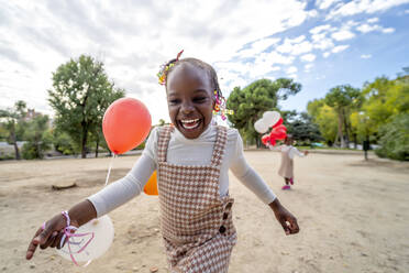 Glückliche afroamerikanische kleine Schwestern in ähnlichen Kleidern stehen mit bunten Luftballons in den Händen auf grünem Gras im Park im Tageslicht - ADSF32321