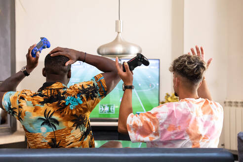 Back view of anonymous multiracial male friends sitting on sofa while playing football videogame on TV in living room - ADSF32364