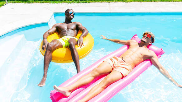 Multirassische männliche Freunde in Badeshorts, die auf einem aufblasbaren Ring und einer Matratze liegen, während sie an einem sonnigen Sommertag ein Sonnenbad im Schwimmbad nehmen - ADSF32365