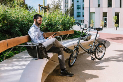 Ganzkörper-Seitenansicht eines männlichen Managers, der auf einer Bank in der Nähe eines Fahrrads sitzt und Nachrichten auf seinem Smartphone im Park liest - ADSF32368