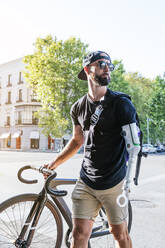 Male hipster with arm prosthesis walking with bicycle on pavement pathway in city - ADSF32372