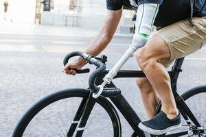 Cropped side view of unrecognizable bearded male hipster with artificial arm riding bicycle along street in summer in city - ADSF32374