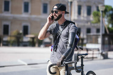 Bearded male with artificial arm walking with bike in street and having conversation on mobile phone on sunny day - ADSF32378