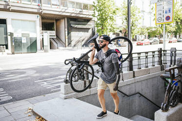 Hoher Winkel eines männlichen Hipsters mit Armprothese, der ein Fahrrad hält und eine Steintreppe in der Stadt hinaufgeht - ADSF32380