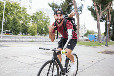 Lustiger männlicher Hipster mit künstlichem Arm, der die Shaka-Geste und die Zunge zeigt, während er mit dem Fahrrad die Straße entlang fährt und in die Kamera schaut - ADSF32381