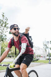 Side view of bearded male hipster with artificial arm riding bicycle along street in summer in city - ADSF32382