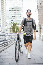 Smiling male hipster with arm prosthesis walking with bicycle on pavement pathway in city looking away - ADSF32383