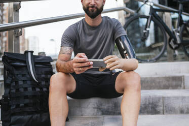 Crop anonymous male with rubber artificial arm browsing mobile phone while sitting on stairs in city - ADSF32384