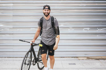 Cheerful male hipster with arm prosthesis standing with bicycle against metal wall in city looking at camera - ADSF32385