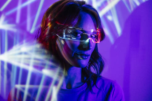 Porträt einer jungen Frau mit Brille und Freizeitkleidung, die in die Kamera schaut, mit leuchtendem Neonlicht im Gesicht und einer futuristischen Bleibrille - ADSF32388
