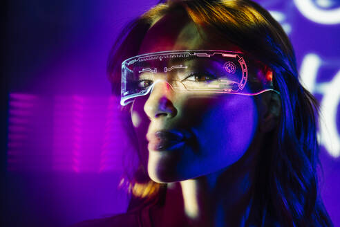 Porträt einer jungen Frau mit Brille und Freizeitkleidung, die in die Kamera schaut, mit leuchtendem Neonlicht im Gesicht und einer futuristischen Bleibrille - ADSF32390