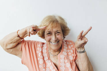 Happy senior woman gesturing peace sign in front of wall - EGHF00265