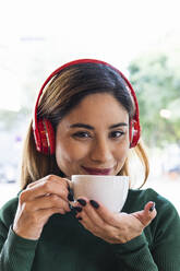 Lächelnde Frau mit kabellosen Kopfhörern und Kaffeetasse im Cafe - PNAF02611