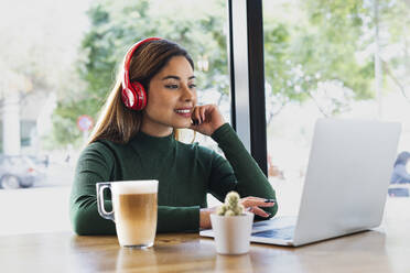 Lächelnde Geschäftsfrau mit Kopfhörern und Tasse bei der Arbeit am Laptop in einem Café - PNAF02612