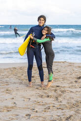 Lächelndes Mädchen mit Kiteboard, das sein Geschwisterchen im Sand am Strand umarmt - DLTSF02461