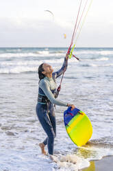 Lächelnde Frau mit Kiteboard und Blick auf den Drachenfallschirm - DLTSF02466