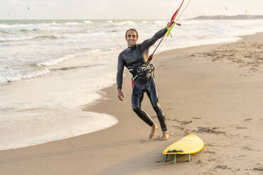 Lächelnder Mann, der das Seil des Fallschirms am Kiteboard am Strand hält - DLTSF02471