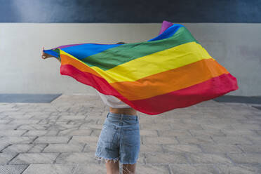 Woman with denim shorts holding rainbow flag on footpath - MGIF01184