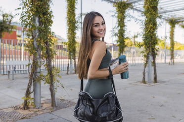 Lächelnder Sportler mit Wasserflasche, der in einem öffentlichen Park über die Schulter schaut - JRVF02134