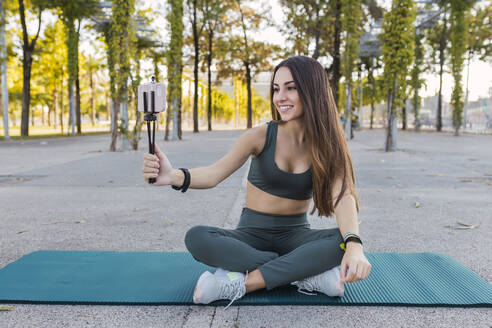 Lächelnde Sportlerin mit Mobiltelefon beim Vloggen auf einer Trainingsmatte im Park - JRVF02136