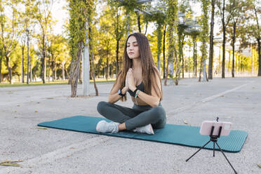 Athlete meditating and vlogging through smart phone at park - JRVF02139