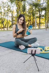 Smiling vlogger pointing at water bottle and filming through mobile phone in park - JRVF02145