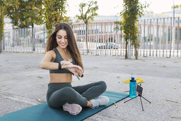 Smiling athlete watching time on exercise mat in public park - JRVF02146