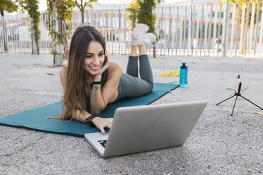 Smiling sportswoman using laptop on exercise mat in public park - JRVF02149