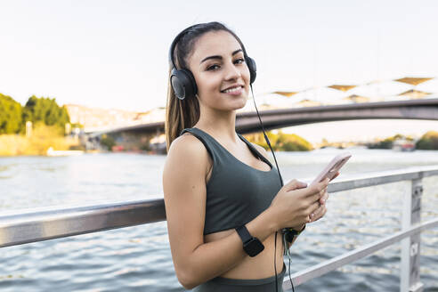 Young athlete with mobile phone listening music through headphones at railing - JRVF02167
