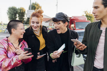 Happy friends showing smart phone to each other on footpath - MEUF05039