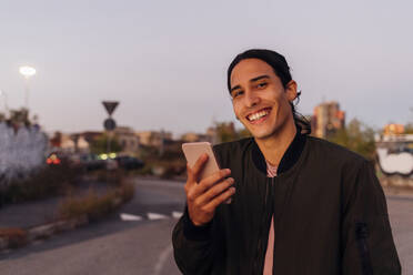 Smiling young man holding smart phone on road - MEUF05064