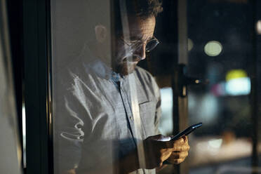 Geschäftsmann, der ein Mobiltelefon benutzt, gesehen durch Glas im Büro bei Nacht - JSRF01756