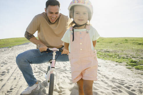 Mädchen mit Helm und Vater auf dem Fahrrad sitzend im Hintergrund - SEAF00187